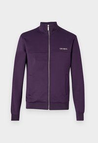 Chaqueta morada con cremallera hecha de tela suave, con cuello alto, dos bolsillos laterales y el texto "LES DEUX" en blanco en el pecho.