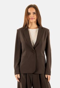 Blazer marrón ajustado en tejido suave, con solapas de muesca, cierre de un solo botón y dos bolsillos frontales. Lleva encima una blusa con estampado.