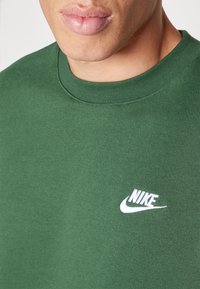 Sudadera de algodón verde con cuello redondo y un logo de Nike en blanco. La tela parece suave con un ligero brillo.