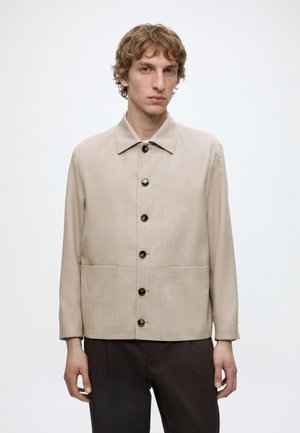 Mladý muž v béžovej zapínanej bunde s golierom a tmavých nohaviciach stojaci pred jednoliatym svetlým pozadím.