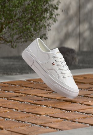 Biały skórzany sneaker z sznurowadłami i małym czerwono-biało-niebieskim logo, unoszący się nad wzorzystą, drewnianą i żwirową powierzchnią na zewnątrz.