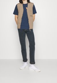 Béžová prošívaná vesta přes tmavě modré tričko s grafikou, v kombinaci s tmavými džínovými kalhotami a bílými teniskami. Jednoduchý design, volný střih.