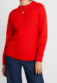 Pull en laine rouge tricoté avec un design texturé, des poignets et un ourlet côtelés, comportant un col rond et des motifs complexes sur la poitrine.