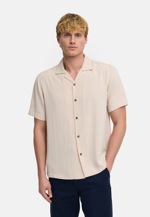 Młody mężczyzna z krótkimi blond włosami, ubrany w beżową koszulę z krótkim rękawem na guziki i ciemne spodnie, stoi z jedną ręką w kieszeni.