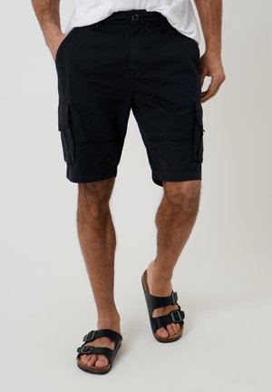 Mann trägt schwarze Cargoshorts und schwarze Sandalen mit doppeltem Riemen und steht mit teilweise in den Taschen befindlichen Händen.