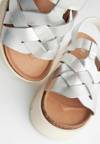 Hõbedased metallilised platvorm sandaalid ristkülikukujuliste rihmadega, pehme pruuni päkkadega ja reljeefse valge tallaga.