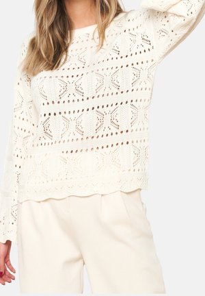 Vrouw draagt een roomkleurige, opengewerkte trui met geometrische patronen en bijpassende roomkleurige broek, tegen een effen achtergrond.