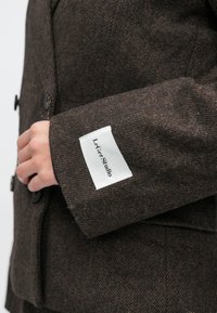 Barna halszálkás blézer texturált gyapjú anyagból, fehér márkajelzéssel az ujján és fekete gombokkal az elején.