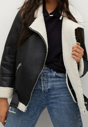 Mujer con chaqueta negra de piel vuelta con forro de forro blanco, suéter oscuro y vaqueros azules, sosteniendo la chaqueta abierta con una mano.