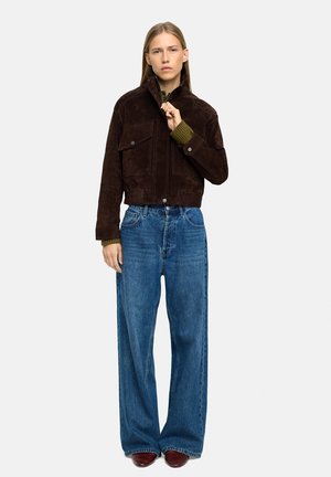 Brązowa zamszowa kurtka cropped z dwiema kieszeniami na piersi, zestawiona z szerokimi niebieskimi dżinsami i bordowymi butami. Prosty i strukturalny design.
