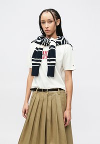 Λευκό μπλουζάκι με κόκκινο κείμενο, μπεζ φούστα με πιέτες και πλεκτό κασκόλ σε ριγέ σχέδιο μπλε σκούρο και άσπρο, περασμένο γύρω από τους ώμους.