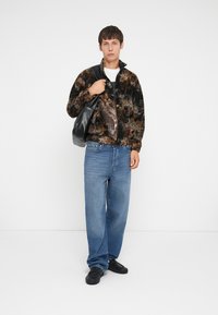 Jachetă din fleece în model abstract maro și negru, asortată cu blugi largi de culoare albastră deschisă, pantofi negri și o geantă de umăr neagră.