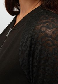 Giacca nera con zip, corpo in tessuto solido e maniche in rete trasparente con stampa leopardata. Il colletto è a coste, aggiungendo texture al design.