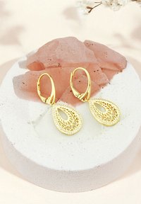 Guldformade tårörhängen har intrikata utskärningar och en kant av små ädelstenar. Visas på en texturerad vit pedestal med rosa kristaller.