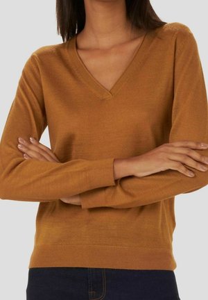 Femme portant un pull marron à col en V avec des manches longues, bras croisés, et un jean bleu foncé, posant devant un fond clair uni.