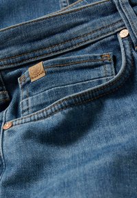 Jeans de mezclilla azul con un bolsillo trasero visible, etiqueta de cuero color marrón claro con un diseño floral y costuras naranjas en contraste.