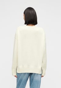 Γυναίκα με ίσια σκούρα μαλλιά, φορώντας ένα υπερμεγέθες κρεμ πουλόβερ και ανοιχτόχρωμο τζιν, με την πλάτη προς τα εμπρός μπροστά σε απλό φόντο.