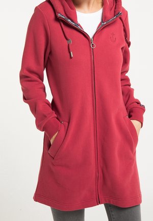 Femme portant un sweat à capuche long rouge avec fermeture éclair, cordons, poches et logo d'ancre sur la poitrine, mains dans les poches, par-dessus une chemise blanche et un pantalon gris.