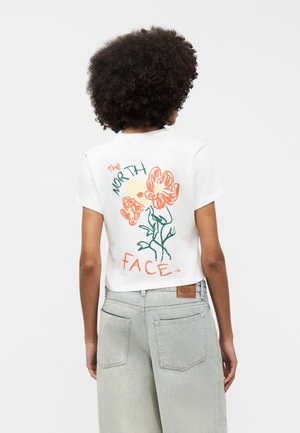 Άτομο με σγουρά μαλλιά που φοράει λευκό κοντομάνικο μπλουζάκι με πορτοκαλί λουλούδια και κείμενο «The North Face» στην πλάτη.