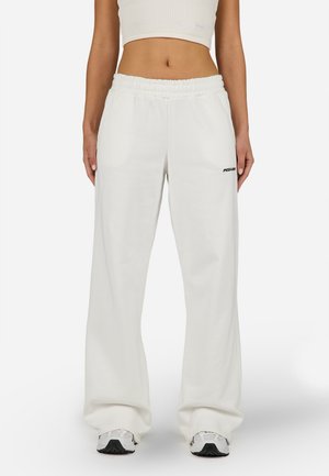 Personne portant un pantalon de survêtement blanc à jambes larges avec une taille élastique et des baskets blanches, debout devant un fond uni.