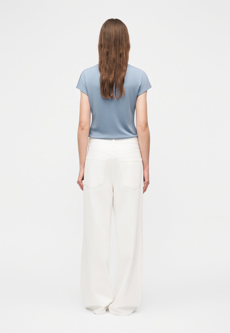 Femme aux longs cheveux bruns portant un haut bleu à manches courtes et un pantalon blanc à jambes larges, debout de dos sur un fond uni.