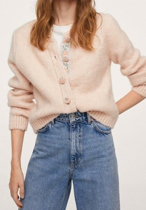 Vrouw draagt een lichtroze, pluizige, gebuttonede cardigan over een witte top, gecombineerd met hooggetailleerde blauwe spijkerbroek, handen deels in de zakken.