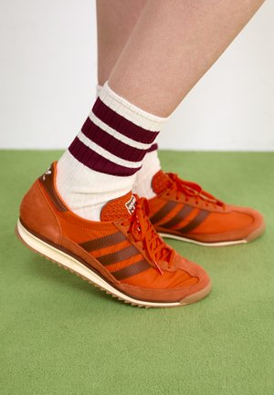 Orange semskede og nylon sneakers med brune striper, røde snørebånd og en teksturert tå. Kombinert med hvite og burgunderstripede ankelsokker.