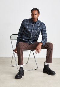 Chemise à carreaux bleu marine et bleu clair, pantalon brun et bottines noires. Chaise en métal plate sur un sol beige texturé.