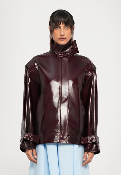 Frau in einer glänzenden dunkelburgunderfarbenen Lederjacke mit hohem Kragen und geknöpften Manschetten, die zu einem hellblauen Rock steht, vor einem weißen Hintergrund.