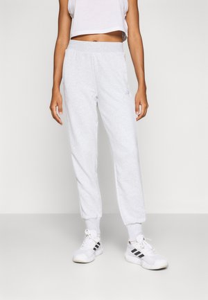 Persoon draagt lichtgrijze joggingbroek, witte crop top, witte sokken en witte sneakers met zwarte strepen, staand tegen een effen achtergrond.