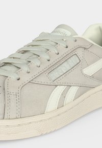 Φωτεινό μπεζ αθλητικό παπούτσι Reebok με λευκές ρίγες, υφασμένο πάτο και κορδόνια, εμφανίζεται σε κοντινό πλάνο από πλάγια γωνία.