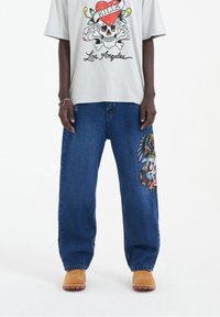 Jasnoszara koszulka z grafiką czaszki, zestawiona z luźnymi niebieskimi dżinsami ozdobionymi kolorowym haftem oraz beżowymi butami.