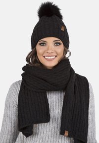 Beanie en tricot noir avec un pompon en fourrure et une écharpe assortie, tous deux présentant un motif texturé. L'écharpe est drapée autour du cou.