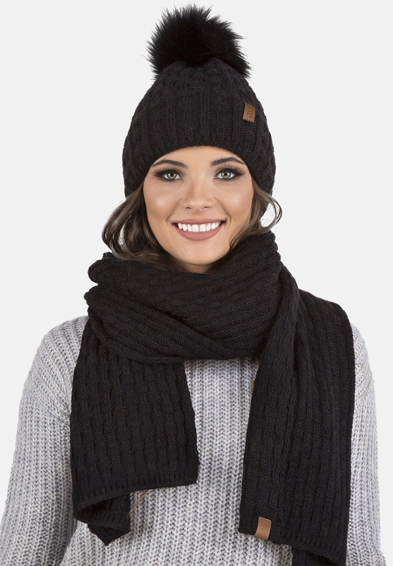 Beanie en tricot noir avec un pompon en fourrure et une écharpe assortie, tous deux présentant un motif texturé. L'écharpe est drapée autour du cou.