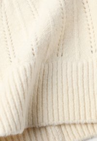 Krémová pletená tkanina s ribovanou textúrou, ktorá obsahuje vertikálne pruhy a dekoratívne otvorené vzory. Mäkký a útulný vzhľad.