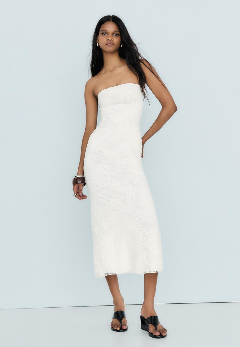 Femme debout contre un mur gris clair portant une robe midi blanche bustier à texture florale et des sandales compensées noires, accessoirisée avec des bracelets.