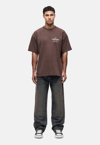 Bruine oversized katoenen t-shirt met witte tekstprint, in combinatie met losse donkere jeans van denim met grote zakken en een vervaagde uitstraling.