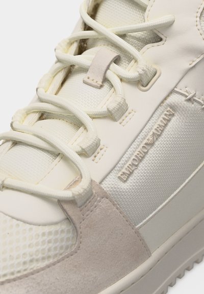 Κοντινό πλάνο άσπρου και μπεζ αθλητικού παπουτσιού με υφασμάτινο πλέγμα υφής, λεπτομέρειες από σουέτ, χοντρές στρογγυλεμένες κορδόνια και το λογότυπο "Emporio Armani" στην πλευρική επιφάνεια.