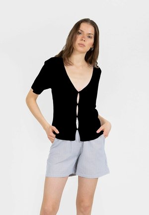 Femme debout, les mains dans les poches, portant un haut noir à manches courtes boutonné et un short rayé bleu clair sur un fond uni.