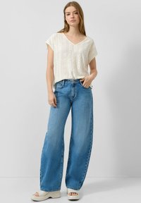 Mujer joven con blusa blanca de manga corta texturizada, vaqueros de pierna ancha azul y sandalias blancas de plataforma, posando frente a un fondo liso.