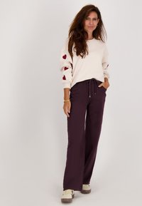 Vrouw die met hand in de zak staat, gekleed in een crèmekleurige sweatshirt met rode harten op de mouwen, donkere wijde broek en beige sneakers.