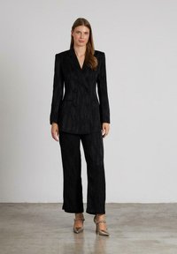 Vrouw draagt een gestructureerde zwarte dubbelgeknopte blazer en bijpassende wijde broek met metallic hoge hakken, terwijl ze op een betonnen vloer staat.