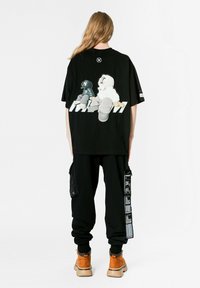 Camiseta negra de gran tamaño con estampado gráfico en la parte trasera, que presenta personajes de dibujos animados. Combinada con pantalones jogger negros y botas marrones.