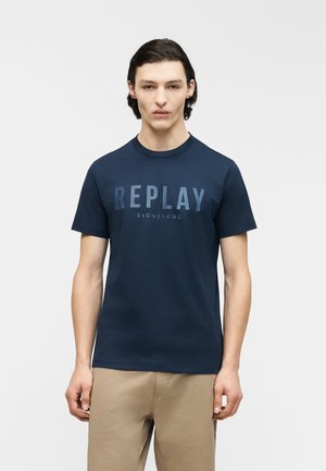 Junger Mann mit dunklem Haar trägt ein marineblaues T-Shirt mit dem Text "REPLAY EIGHTYONE" und beigefarbene Hose, steht vor einem schlichten hellen Hintergrund.