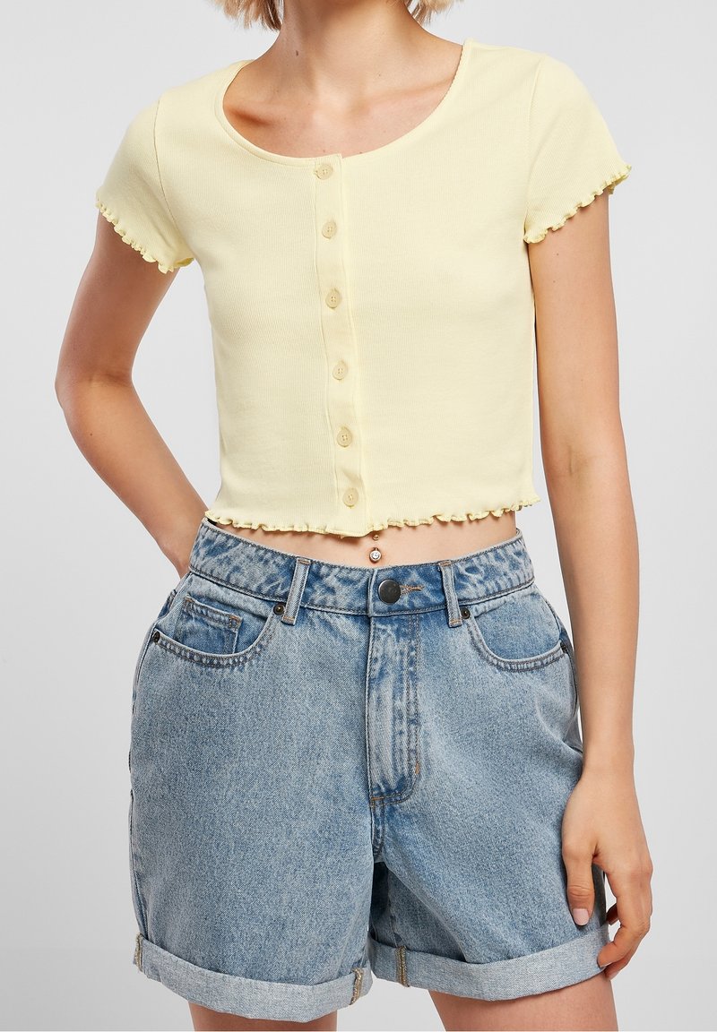 Femme portant un crop top boutonné à manches courtes jaune clair et un short en denim bleu clair à taille haute avec revers, debout avec une main dans la poche.