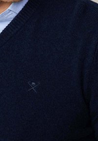 Homme portant un pull en maille bleu foncé avec un petit logo brodé représentant une balle de golf et des clubs croisés sur la poitrine, par-dessus une chemise bleu clair.