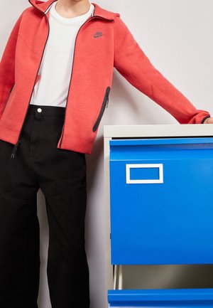 Personne portant une veste zippée rouge Nike et un pantalon noir, debout avec un bras reposant sur un classeur bleu.