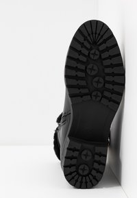 Botte noire avec une semelle en caoutchouc texturée, dotée de crampons profonds et d'une bordure en fausse fourrure. Le design est fonctionnel avec une fermeture éclair latérale.