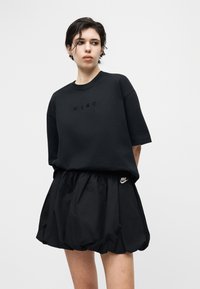 Νέος/νέα με μαύρο υπερμεγέθες μπλουζάκι Nike και μαύρη φούστα μπαλόνι, με γκρι απλό φόντο, κοιτάζοντας προς τα πλάγια.