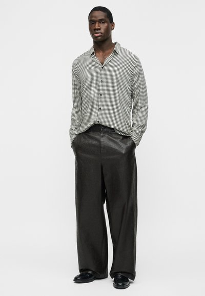 Homem vestindo camisa de manga comprida xadrez preta e branca, enfiada em calças largas de couro pretas, com as mãos nos bolsos, em pé contra um fundo branco.